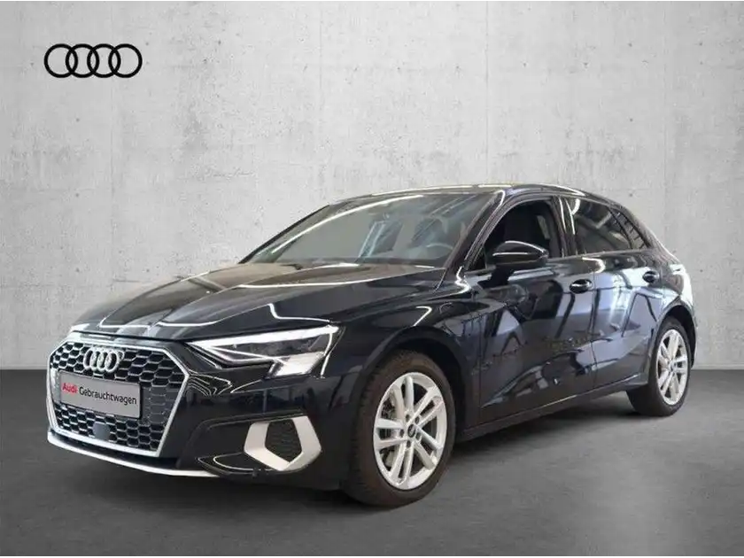 Das Bild zeigt einen schwarzen AUDI A3 SPORTBACK von der Seite. Im Hintergrund ist ein grauer Wandanstrich zu sehen, und es gibt ein Händlerlogo oben im Bild.