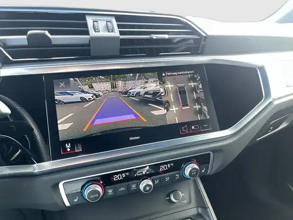 Das Bild zeigt den Innenraum eines AUDI Q3 mit einem Bildschirm, der eine Rückfahrkameraansicht anzeigt. Die Interieurfarbe ist schwarz und es sind markante Texteinblendungen auf dem Bildschirm vorhanden.