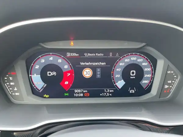 Das Bild zeigt das Cockpit eines AUDI Q3. Es handelt sich um eine Innenaufnahme mit einem digitalen Armaturenbrett in schwarzer Farbe und markanten Texteinblendungen, einschließlich Verkehrsschildern.