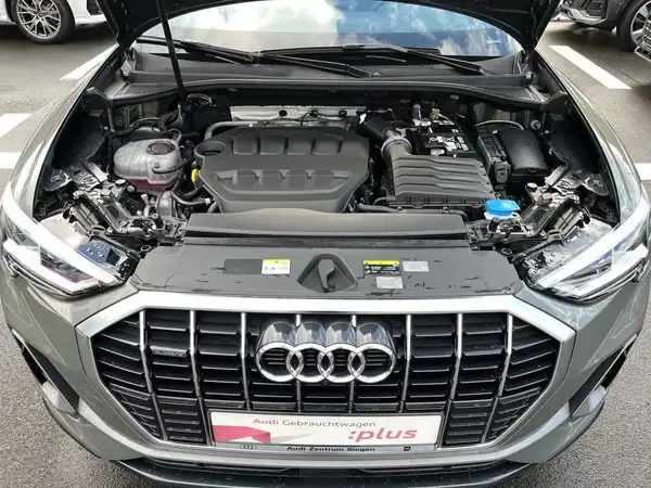 Das Bild zeigt den Motorraum eines Audi Q3, aufgenommen von oben. Das Fahrzeug ist in einer grauen Farbe, wobei das Händlerlogo im Bild sichtbar ist und es einige markante Texteinblendungen gibt.