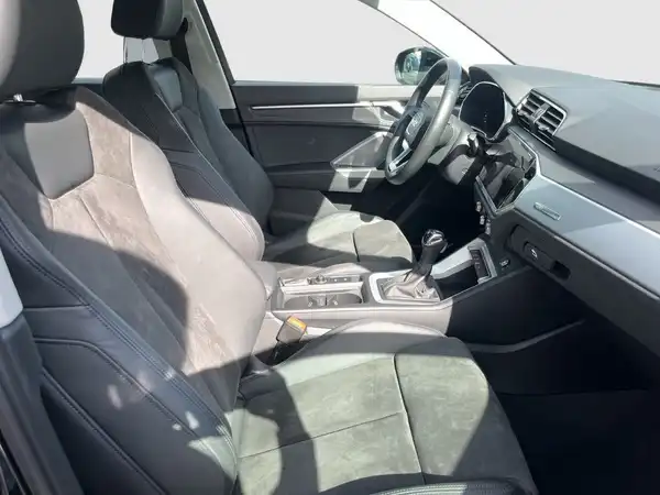 Das Bild zeigt den Innenraum eines AUDI Q3, gesehen aus der Fahrerseite. Die Sitze sind grau und bestehen aus einer Kombination aus Stoff und Leder.