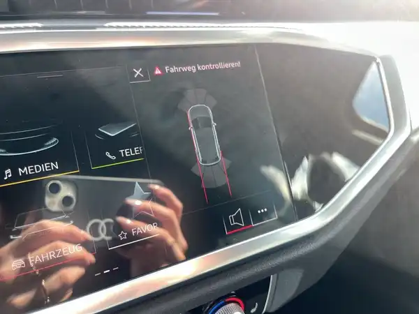 Das Bild zeigt das Innere eines AUDI Q3 mit einem Blick auf das Navigationsdisplay. Die Interieurfarbe ist schwarz. Es sind markante Texteinblendungen sichtbar.