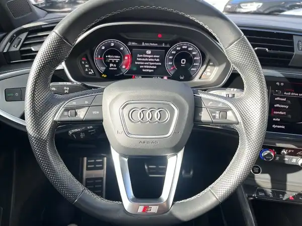 Innenaufnahme des Fahrzeugs Audi Q3, zeigt das Lenkrad und das Dashboard in schwarzem Leder.
