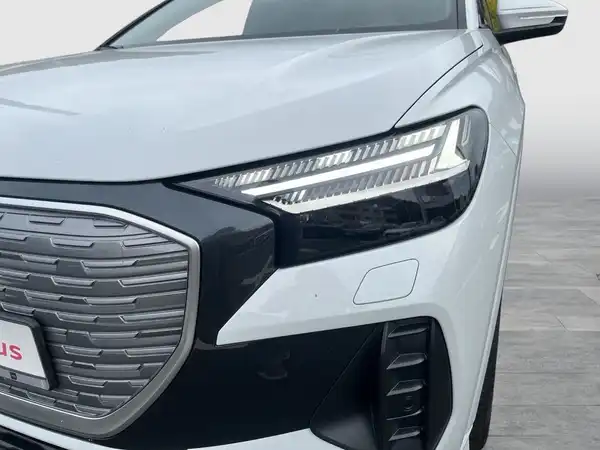 Das Bild zeigt einen weißen AUDI Q4 E-TRON aus der Frontalansicht. Es enthält das Händlerlogo und andere markante Texteinblendungen.
