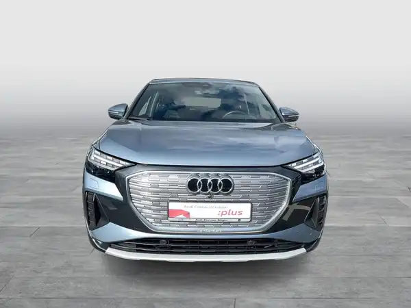 Das Bild zeigt einen grauen AUDI Q4 SPORTBACK E-TRON aus der Frontalansicht. Ein Händlerlogo ist auf dem Bild zu sehen, und es enthält markante Texteinblendungen.