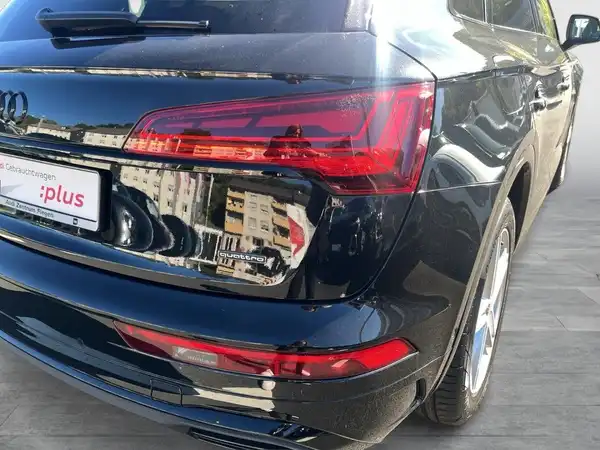 Das Bild zeigt die Rückansicht eines schwarzen AUDI Q5. Im Hintergrund sind Gebäudespiegelungen sichtbar. Das Händlerlogo ist eingeblendet.
