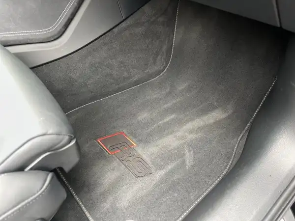 Das Bild zeigt den Innenraum eines Fahrzeugs, spezifisch den Fußraum auf der Fahrerseite mit einem schwarzen Teppich und einem roten Logo 'S'.