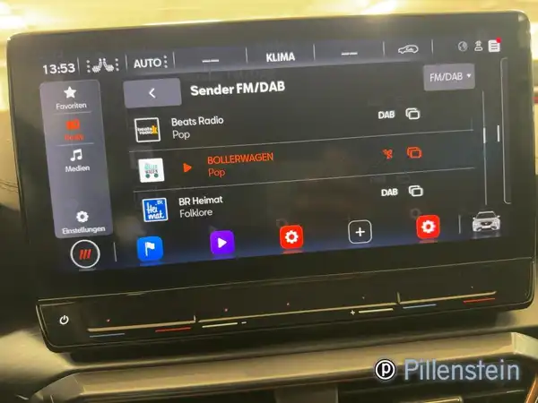 Das Bild zeigt das Innenleben eines Fahrzeugs mit einem Bildschirm, der Radio- und Klimafunktionen anzeigt. Es ist ein schwarzes Interieur zu sehen, und das Händlerlogo ist sichtbar.