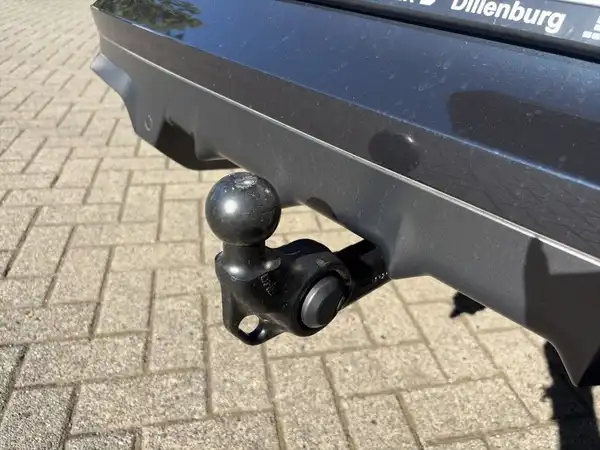 Das Bild zeigt die Rückansicht eines schwarzen Fahrzeugs mit einer Anhängerkupplung, das auf einem gepflasterten Weg steht. Ein Händlerlogo ist sichtbar.