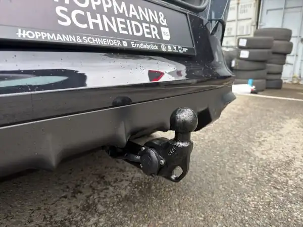 Das Bild zeigt die Rückseite eines schwarzen Fahrzeugs mit einem sichtbaren Anhängerkupplungsmodul. Im Hintergrund sind einige Autoreifen zu sehen und ein Händlerlogo ist eingeblendet.