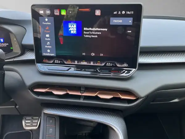 Das Bild zeigt das Innere eines Fahrzeugs, mit einem schwarzen Interieur und einem großen digitalen Bildschirm, der Musikinformationen anzeigt.
