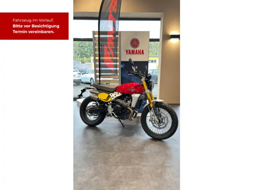 Das Bild zeigt ein FANTIC CABALLERO 500 Motorrad, aufgenommen von der Seite. Es hat eine rote, schwarze und gelbe Farbkombination. Im Hintergrund sind Händlerlogos und eine Texteinblendung sichtbar.