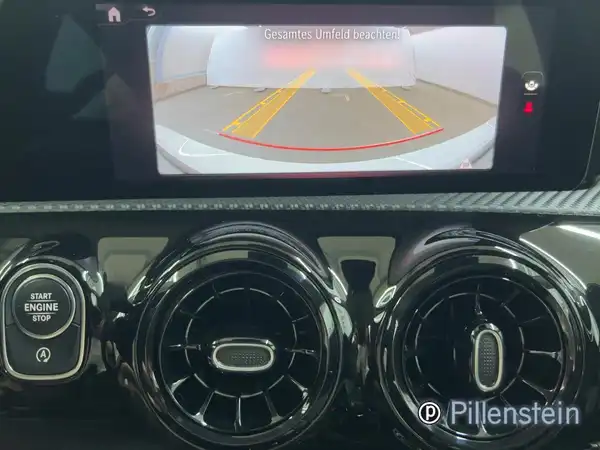Das Bild zeigt das Innenleben eines Mercedes-Benz A 180 d, einschließlich des Navigationsbildschirms mit Rückfahrkameraansicht und der Klimaanlagenbedienelemente in schwarzem Interieur.