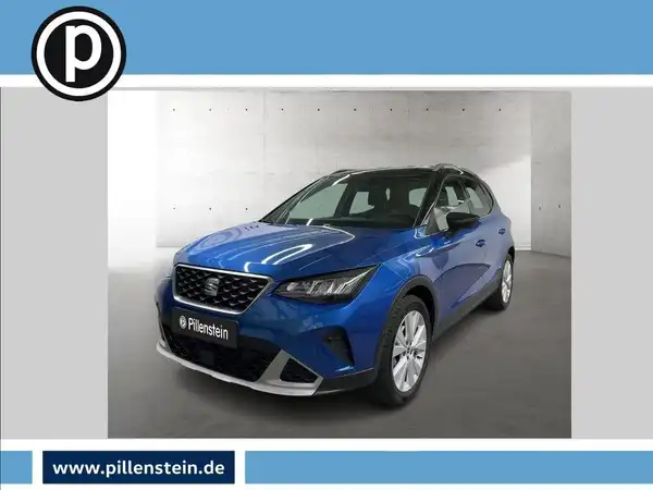 Das Bild zeigt einen blauen SEAT Arona von vorne, aufgenommen in einem Studio mit neutralem Hintergrund. Es ist das Händlerlogo sichtbar.