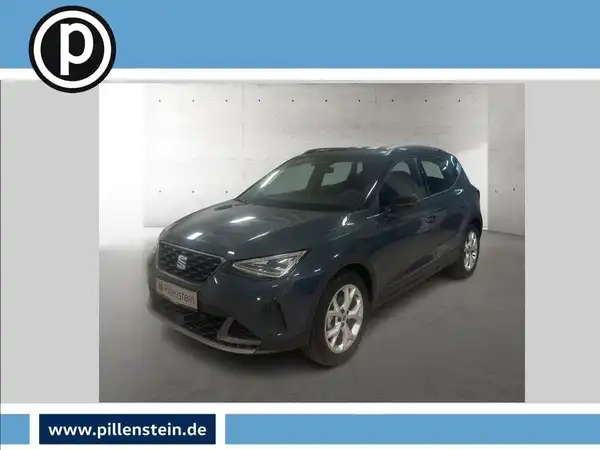 Das Bild zeigt einen SEAT ARONA, aufgenommen von der Seite in dunkelgrauer Farbe. Das Händlerslogo ist eingeblendet und das Foto enthält markante Texteinblendungen.