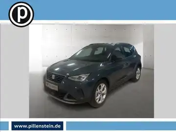 Das Bild zeigt einen SEAT ARONA, aufgenommen von der Seite in dunkelgrauer Farbe. Das Händlerslogo ist eingeblendet und das Foto enthält markante Texteinblendungen.