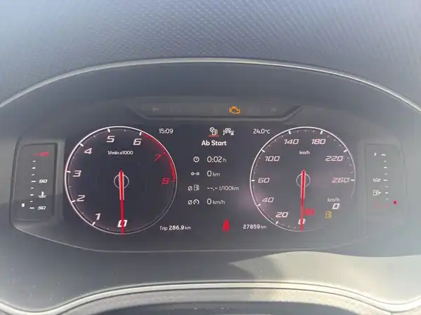 Das Bild zeigt das Armaturenbrett eines Fahrzeugs mit digitalem Display und Instrumenten. Es handelt sich um eine Innenaufnahme des Fahrzeugs, das Interieur ist schwarz und es sind markante Texteinblendungen vorhanden.