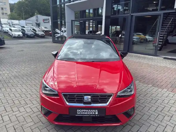 Das Bild zeigt einen roten SEAT IBIZA, aufgenommen von vorne vor einem Autohaus. Es ist ein Händlerlogo sichtbar.