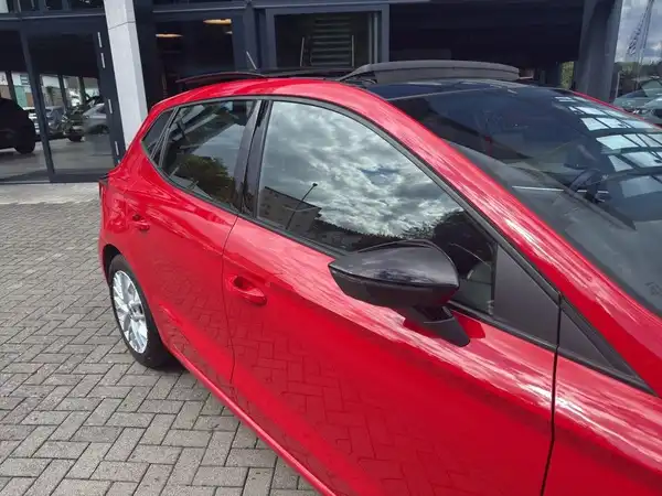 Das Bild zeigt einen roten SEAT IBIZA von der Seite, aufgenommen auf einem Parkplatz mit einem Autohaus im Hintergrund.