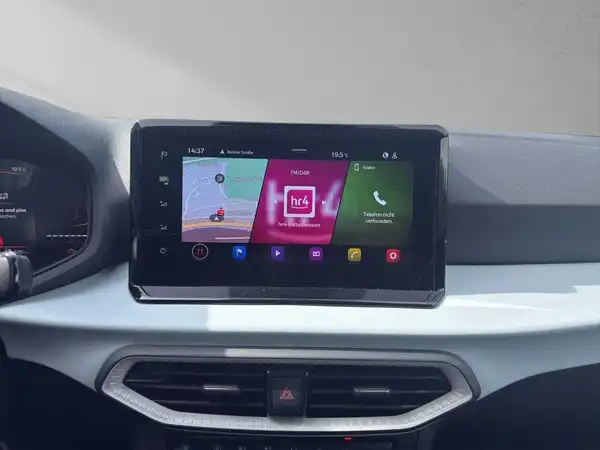 Das Bild zeigt das Innere eines Fahrzeugs mit einem Bildschirm für Navigation und Unterhaltung. Es sind einige Apps und Funktionen sichtbar, das Interieur ist schwarz mit silbernen Akzenten.