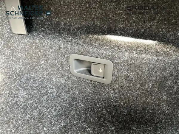 Das Bild zeigt einen Blick ins Innere des ŠKODA ENYAQ iV, speziell auf einen Teil des Kofferraums mit einem grauen Teppichboden und einem Bedientaster. Das Händlerlogo ist sichtbar.