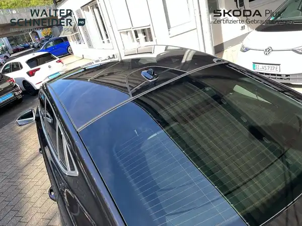 Das Bild zeigt einen schwarzen ŠKODA ENYAQ iV aus der Seitenansicht. Im Hintergrund sind andere Fahrzeuge und Teile des Autohauses erkennbar. Das Händlertlogo ist im Bild sichtbar, außerdem sind markante Texteinblendungen vorhanden.