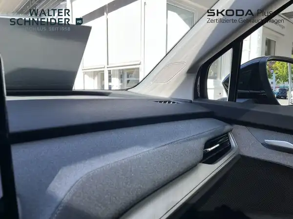 Das Bild zeigt das Innere eines ŠKODA ENYAQ iV mit einem schwarzen Armaturenbrett und einer Seitenansicht des Innenraums. Es enthält das Händlerinformationen.