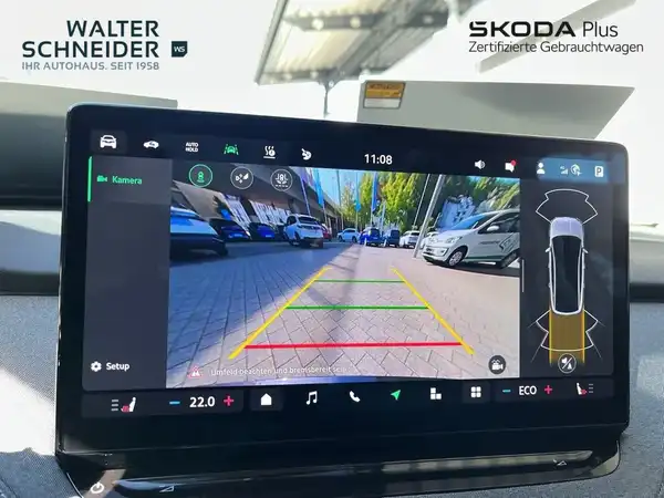 Das Bild zeigt das Innenleben eines ŠKODA ENYAQ iV mit einem auf dem Bildschirm angezeigten Rückfahrkamerabild. Der Innenraum ist schwarz und enthält das Händlerlogo sowie weitere Texteinblendungen.