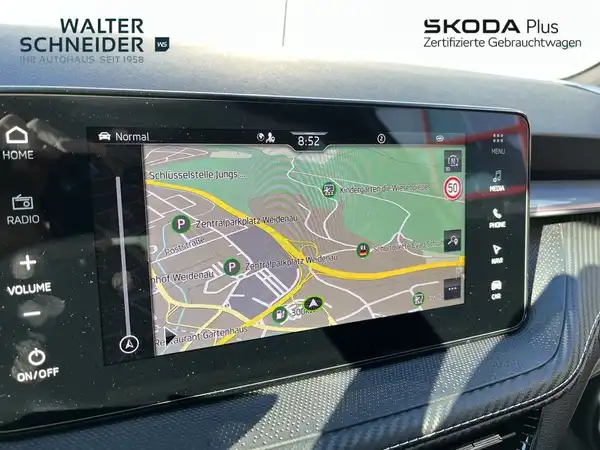 Das Bild zeigt den Innenraum eines ŠKODA Fahrzeugs mit einem Navigationsdisplay, das eine Karte anzeigt. Das Interieur ist modern und überwiegend schwarz. Das Händlerlogo ist sichtbar.