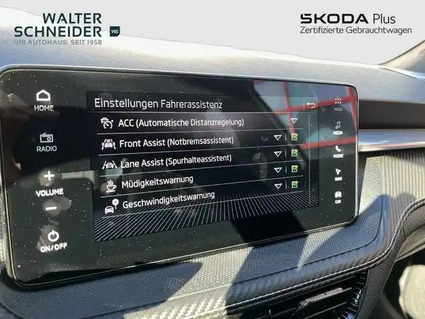 Das Bild zeigt das Innenraumdisplay eines ŠKODA FABIAs mit schwarz gefärbtem Interieur. Es enthält verschiedene Einstellungen zur Fahrerassistenz. Das Händlerlogo ist sichtbar.