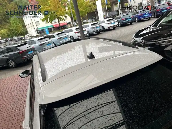 Das Bild zeigt einen weißen ŠKODA KAMIQ von der Seite in einem Autohausparkplatz, mit sichtbaren Händlerlogos und anderen Fahrzeugen im Hintergrund.