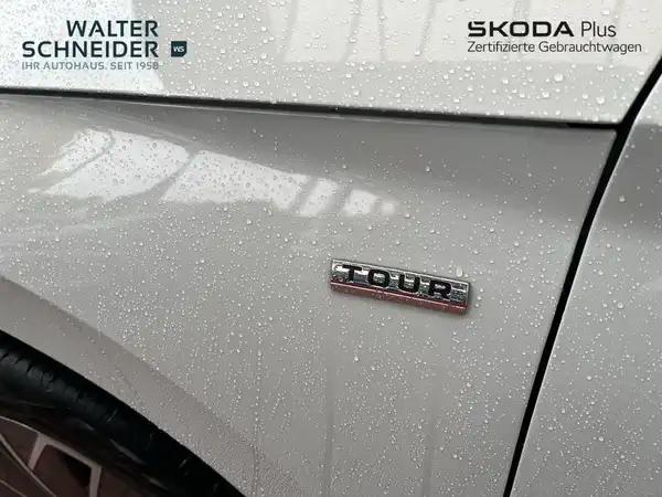 Das Bild zeigt die Seite eines weißes ŠKODA KAMIQ mit Regentropfen darauf. Auf dem Fahrzeug ist ein Badge mit der Bezeichnung 'TOUR' zu sehen. Im oberen Bereich des Bildes sind die Logos und Kontaktdaten des Händlers Walter Schneider eingeblendet.