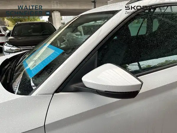 Das Bild zeigt die seitliche Ansicht eines weißen ŠKODA KAMIQ. Es sind Händlerlogos und markante Texteinblendungen sichtbar.