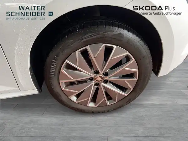Das Bild zeigt das Rad eines weißen ŠKODA KAMIQ von der Seite. Im Hintergrund sind Händlerlogos und Textinformationen sichtbar.