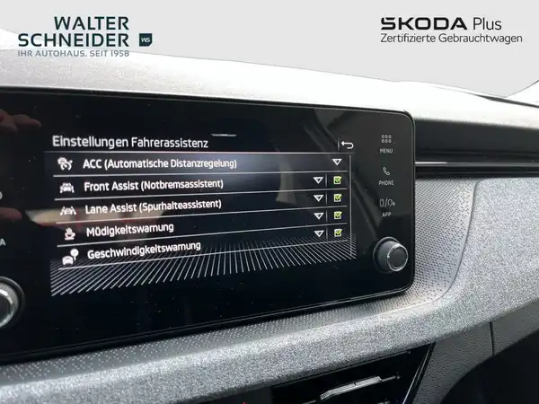 Das Bild zeigt das Innere eines ŠKODA KAMIQ mit einem Fokus auf das Navigationsdisplay, das Einstellungen für die Fahrerassistenz anzeigt. Die Innenfarbe ist schwarz und das Händlerlogo ist sichtbar.