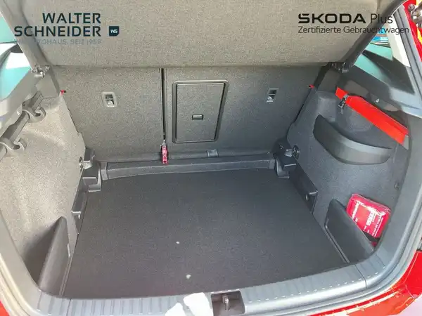 Das Bild zeigt den Innenraum des Kofferraums eines roten ŠKODA KAROQ. Das Fahrzeug hat schwarze Interieurelemente. Zusätzlich sind Händlerlogos eingeblendet.