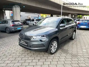 Das Bild zeigt einen ŠKODA KAROQ aus der Seitenansicht. Das Fahrzeug ist grau und es ist ein Händlerlogo eingeblendet, zusätzlich gibt es markante Texteinblendungen.