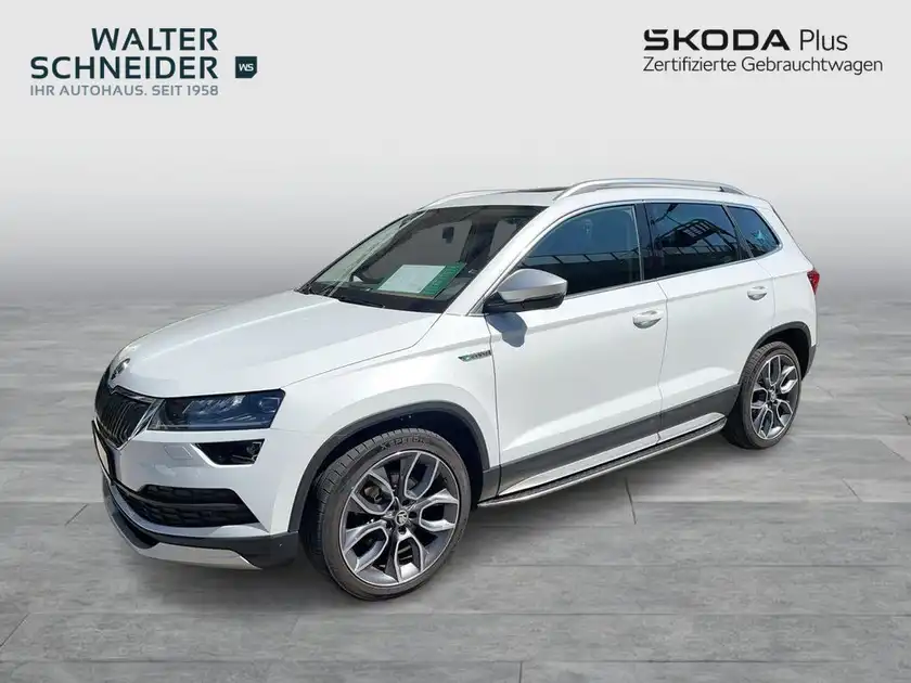 Das Bild zeigt einen weißen ŠKODA KAROQ aus der Seitenansicht. Im Hintergrund sind die Logos des Händlers zu sehen.