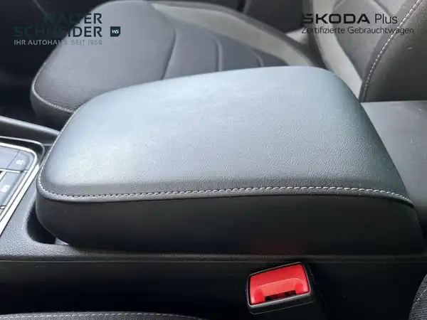 Das Bild zeigt den Innenraum eines ŠKODA KODIAQ mit einem schwarzen Armaturenbrett und einem mittleren Armlehnenbereich. Es gibt ein Händlerlogo im Bild.