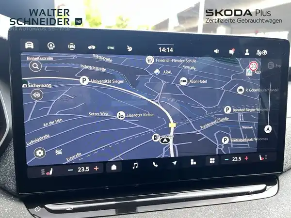 Das Bild zeigt den Innenraum eines ŠKODA-Fahrzeugs mit einem Infotainment-Bildschirm, auf dem eine Navigationskarte angezeigt wird. Das Interieur ist schwarz. Ein Händlerlogo ist im Bild vorhanden und es gibt markante Texteinblendungen.