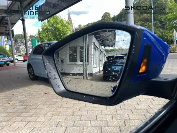 Das Bild zeigt einen blauen ŠKODA OCTAVIA von der Seite, aufgenommen durch den linken Außenspiegel. Im Hintergrund sind mehrere Fahrzeuge und das Autohaus sichtbar, dabei sind andere Fahrzeuge unscharf zu erkennen.