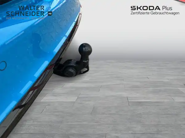 Das Bild zeigt einen hinteren Blick auf ein blaues Fahrzeug, wahrscheinlich einen ŠKODA, mit einem sichtbaren Anhängerkupplung. Auf dem Bild sind ein Händlerlogo und markante Texteinblendungen zu sehen.
