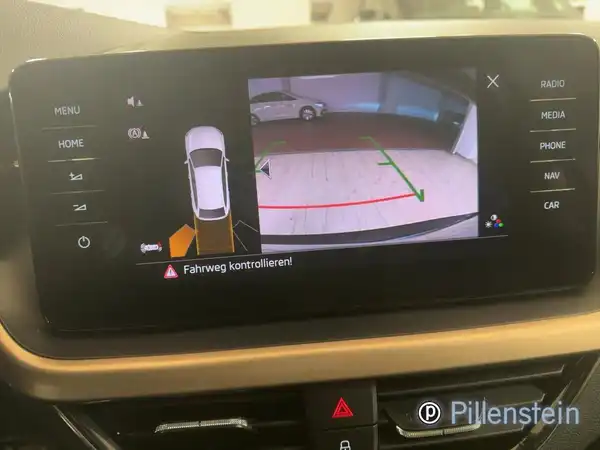 Das Bild zeigt das Innenleben eines Fahrzeugs, speziell den Bildschirm des Navigations- oder Informationssystems mit einer Rückfahrkameraansicht. Die Inneneinrichtung ist dunkel gestaltet. Das Händlerlogo ist abgebildet. Es gibt markante Texteinblendungen auf dem Bildschirm.