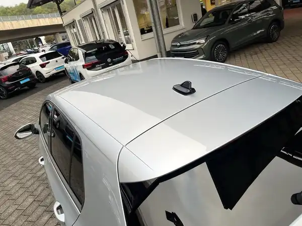 Das Bild zeigt einen VW Golf aus der Seitenansicht, in silberner Farbe, auf einem Parkplatz mit weiteren Fahrzeugen im Hintergrund.