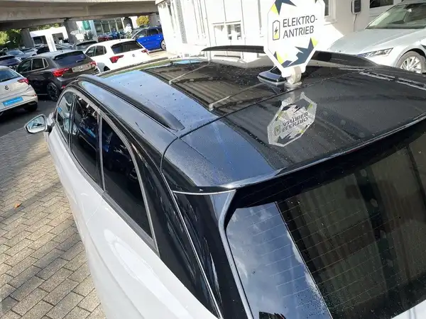 Das Bild zeigt einen weißen VW ID.7, aufgenommen von hinten mit einem Händlerlogo und einem Text 'ELEKTRO-ANTRIEB' auf dem Dach. Das Fahrzeug steht auf einem Parkplatz.