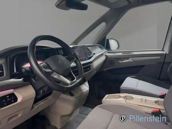 Das Bild zeigt das Innere eines VW Multivan, aufgenommen aus der Perspektive des Fahrers. Das Interieur ist in beigem und grauem Farbton gehalten, und es enthält das Händlerlogo.