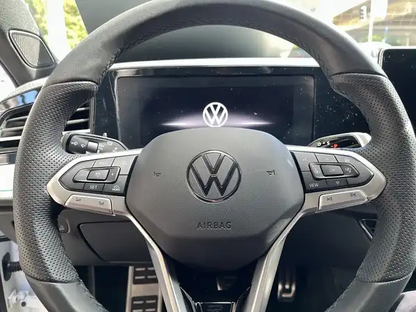 Das Bild zeigt das Innere eines VW Passat, insbesondere das Lenkrad und das Armaturenbrett. Das Lenkrad ist mit einer schwarzen Ledertextur ausgestattet und es ist das VW-Logo sichtbar.