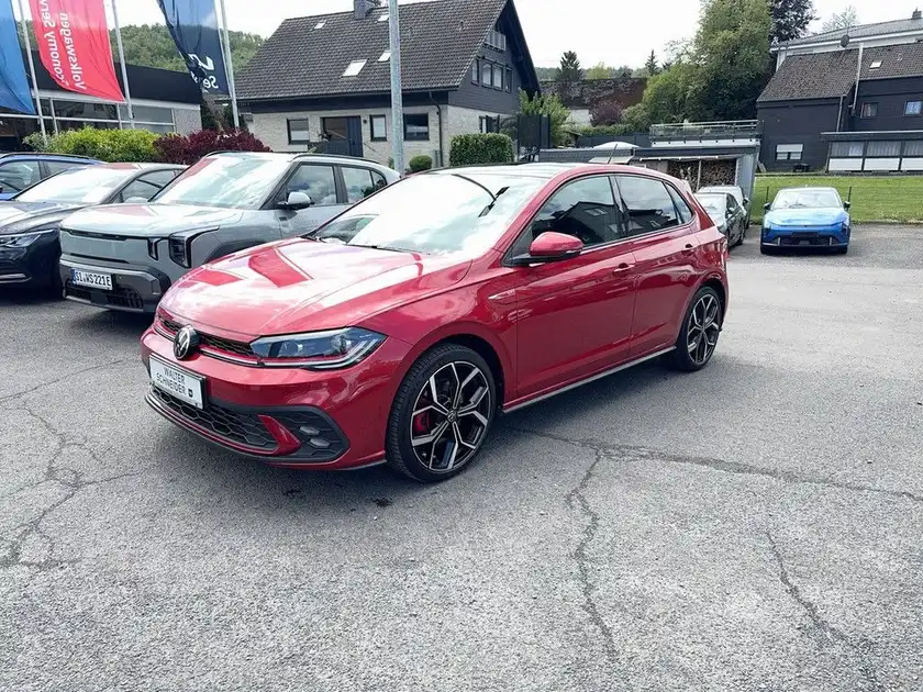 Das Bild zeigt einen roten VW Polo GTI von der Seite. Im Hintergrund sind weitere Fahrzeuge und ein Händlerlogo sichtbar.