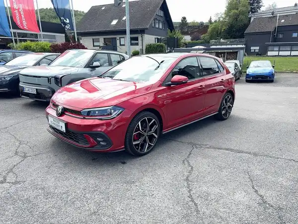 Das Bild zeigt einen roten VW Polo GTI von der Seite. Im Hintergrund sind weitere Fahrzeuge und ein Händlerlogo sichtbar.