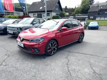 Das Bild zeigt einen roten VW Polo GTI von der Seite. Im Hintergrund sind weitere Fahrzeuge und ein Händlerlogo sichtbar.